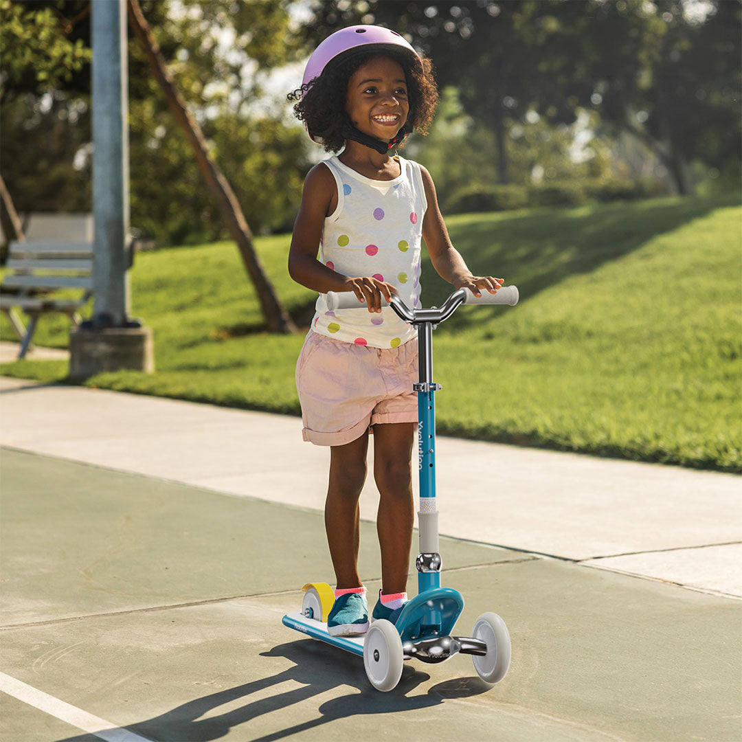 Een lachend kind met roze helm rijdt buiten op de blauwe Yvolution kinderstep.