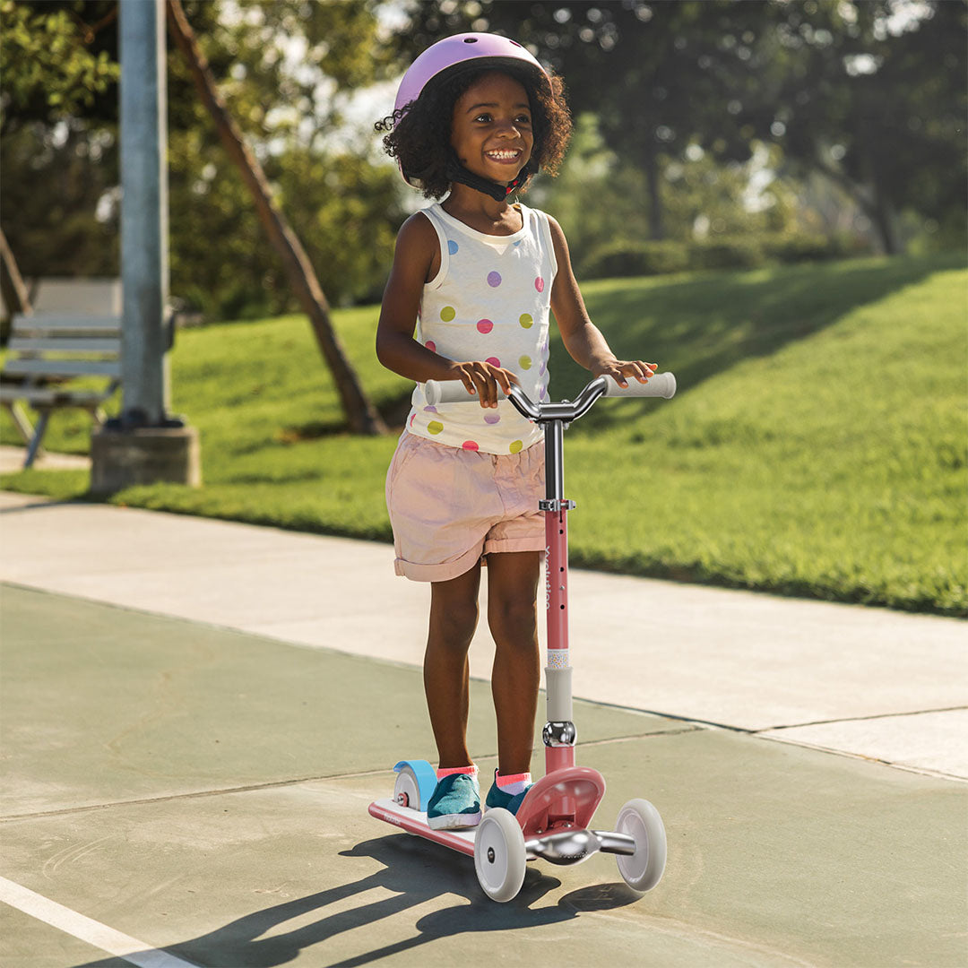 Een lachend kind met roze helm rijdt buiten op de rode Yvolution kinderstep.
