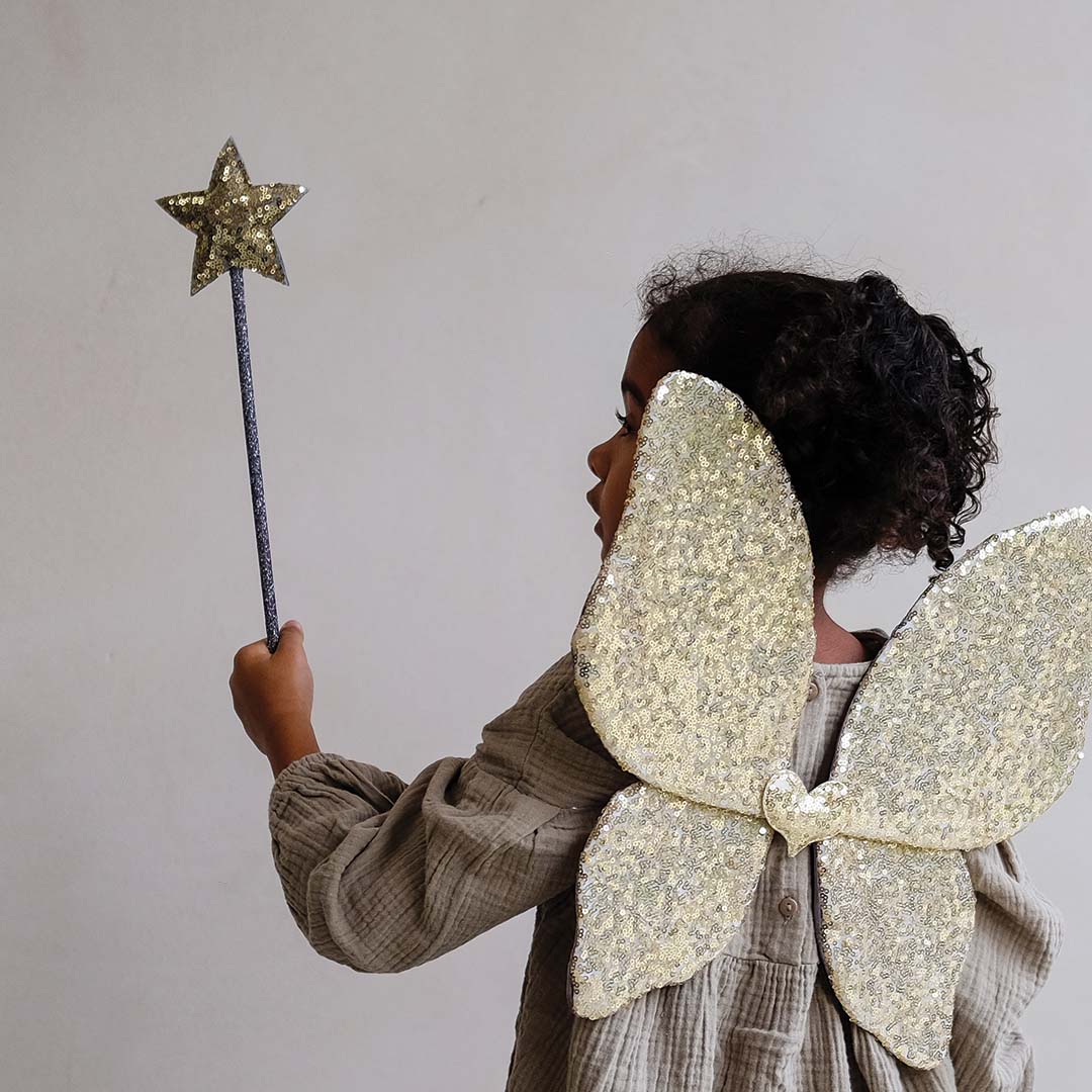 Meisje met gouden pailletten vlindervleugels houdt een mimi & lula toverstaf met gouden ster omhoog, gezien van achteren.