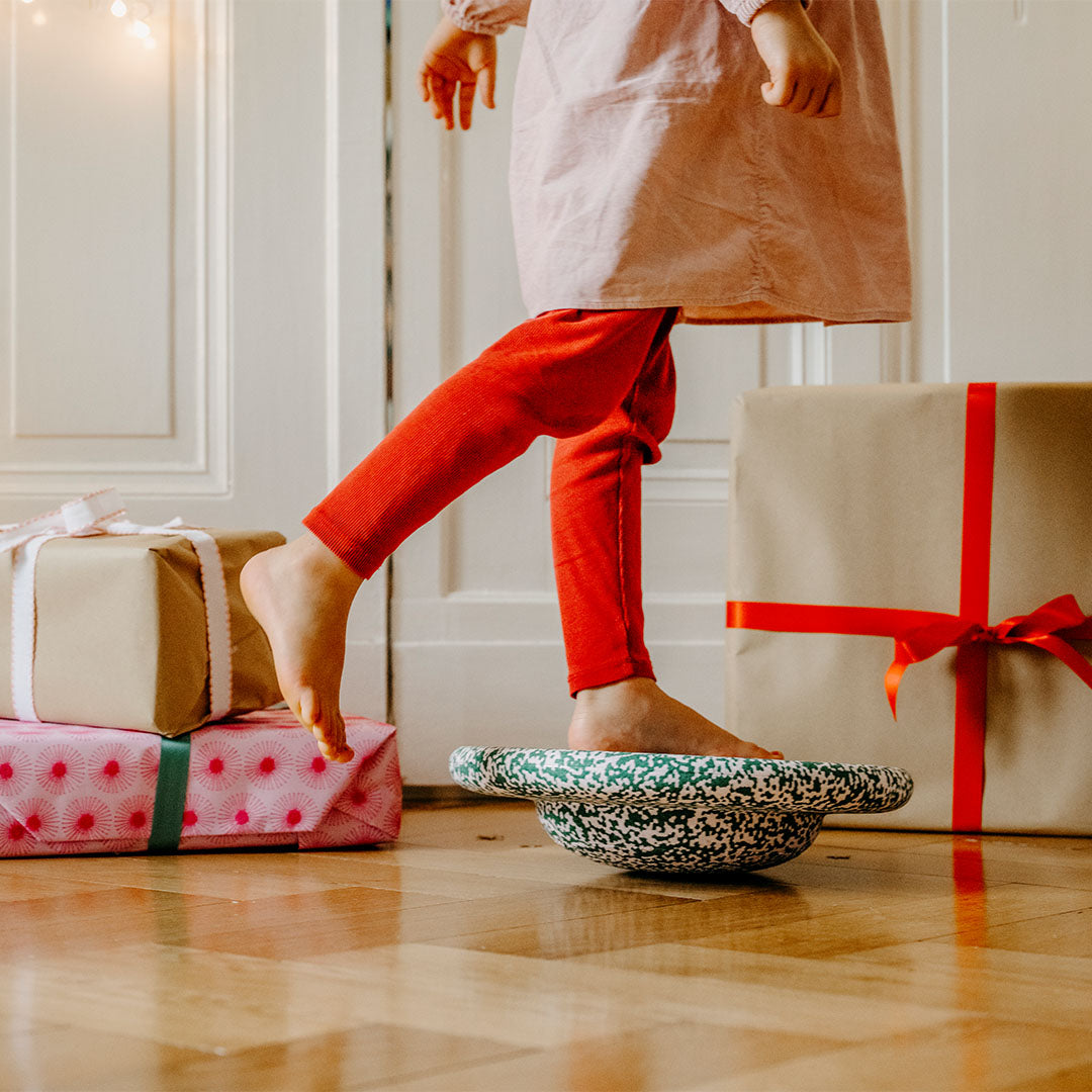 Een kind balanceert met blote voeten op een Stapelstein balance board, met cadeaus op de achtergrond.