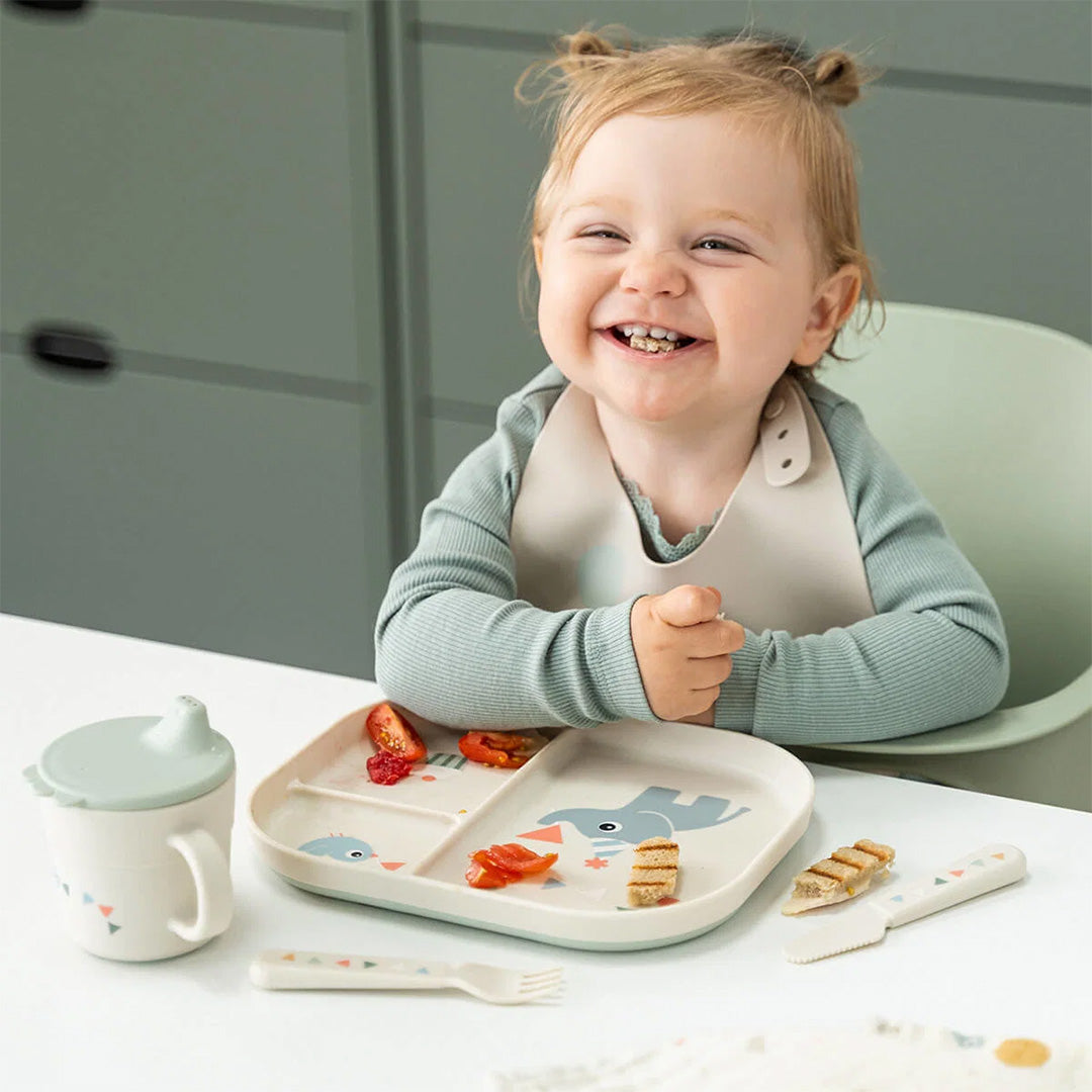 Een lachende baby zit aan tafel met een Done by Deer voedingsset, inclusief het kinderbestek en een bord met olifant en vogel.