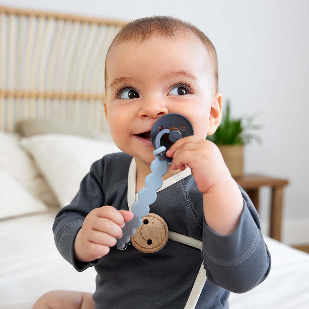 Een baby met een donkergrijze romper draagt het lässig speenkoord met een donkergrijze speen.