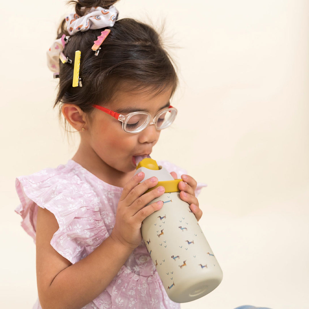 Een meisje met een bril drinkt uit een beige drinkfles met een patroon van teckels.