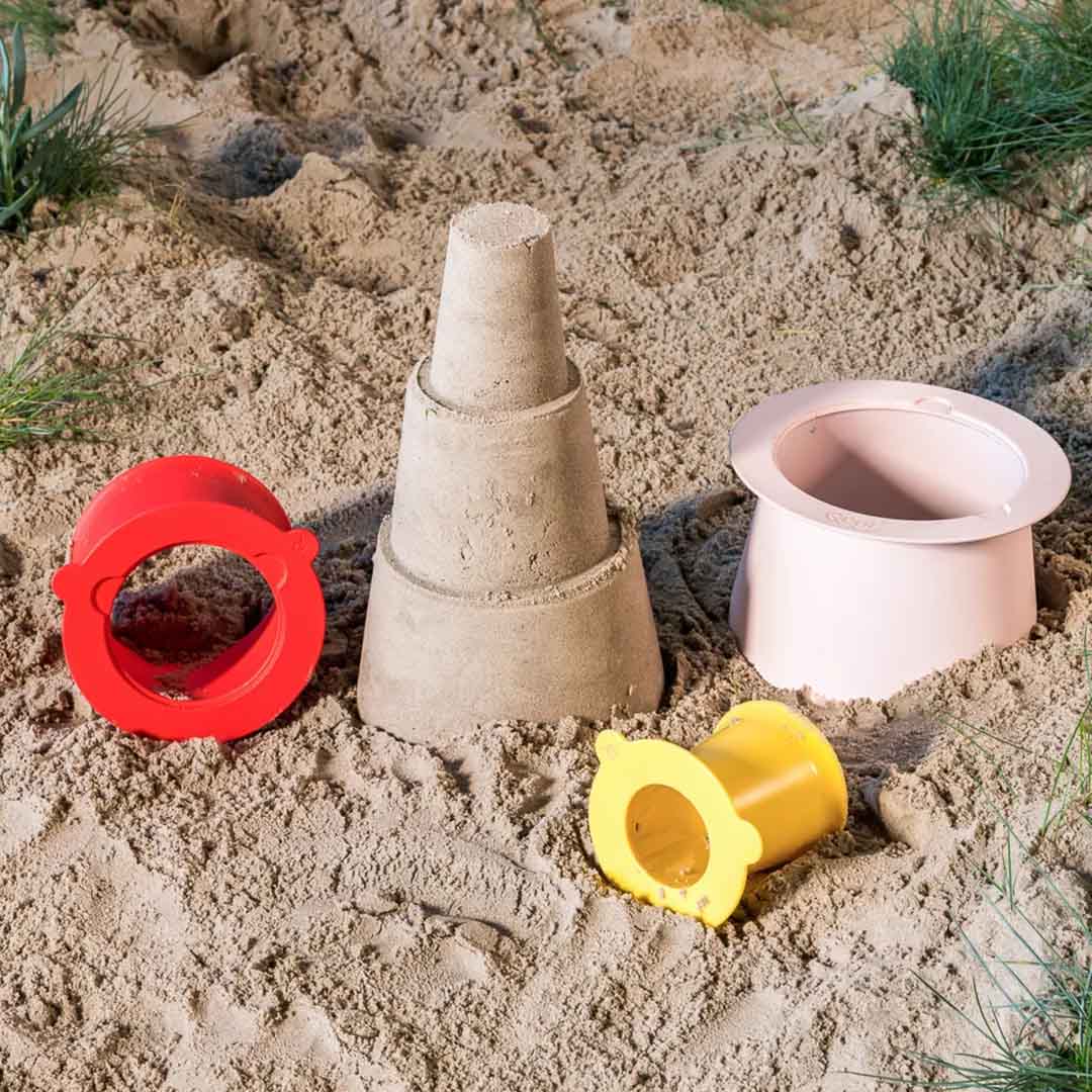 quut zandstapeltoren onderdelen in rood, geel en roze met een zandkasteel in het zand.