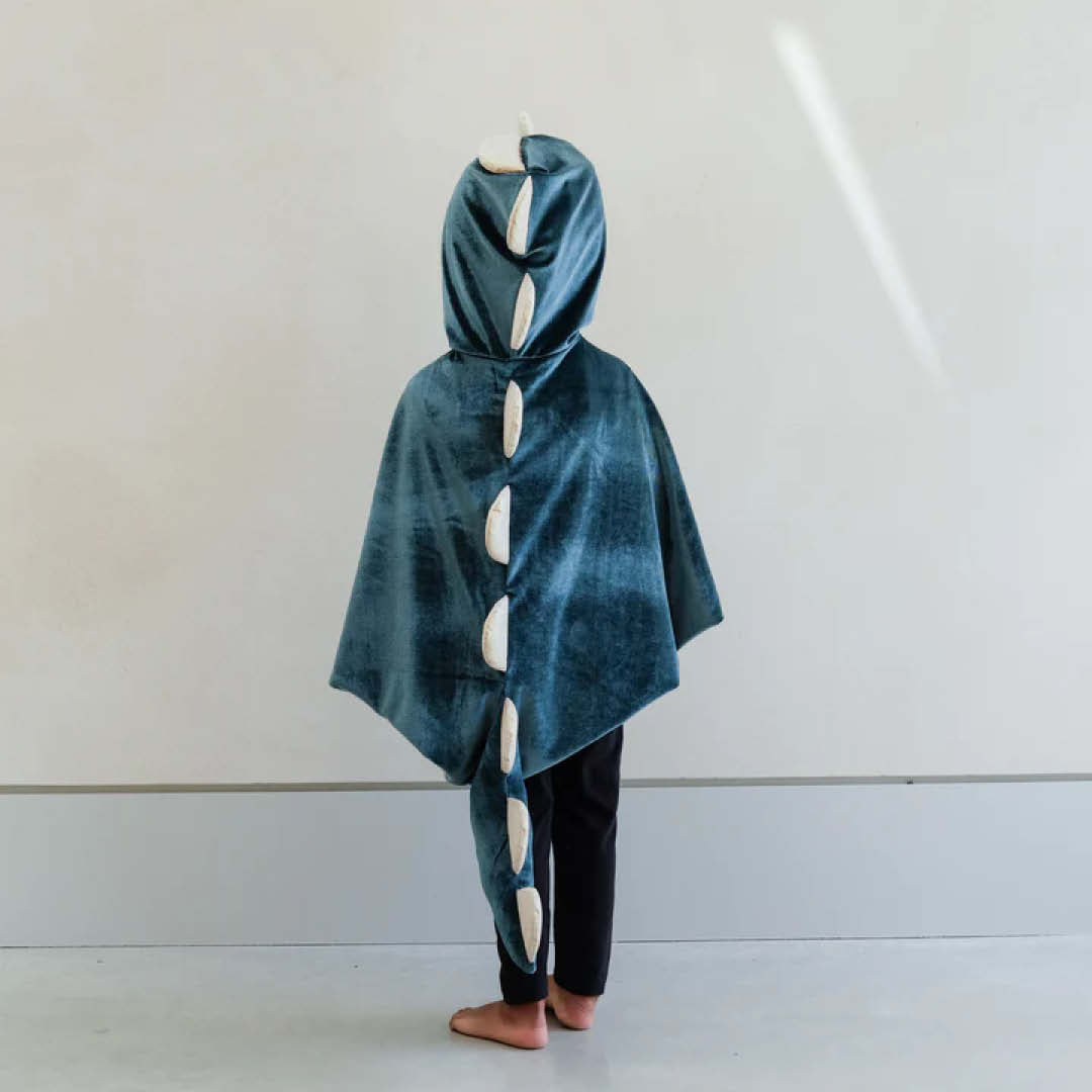 Kind draagt de Mimi & Lula groene fluwelen dinosaurus verkleedcape, van achteren gefotografeerd, met de staart en stekels duidelijk zichtbaar.