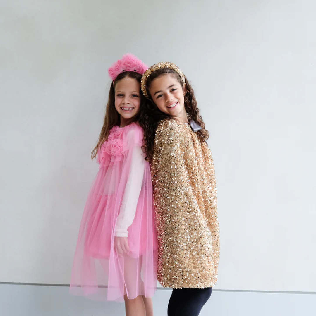 Twee lachende meisjes, waarvan één de gouden pailletten verkleedcape van Mimi & Lula draagt, staan rug aan rug.