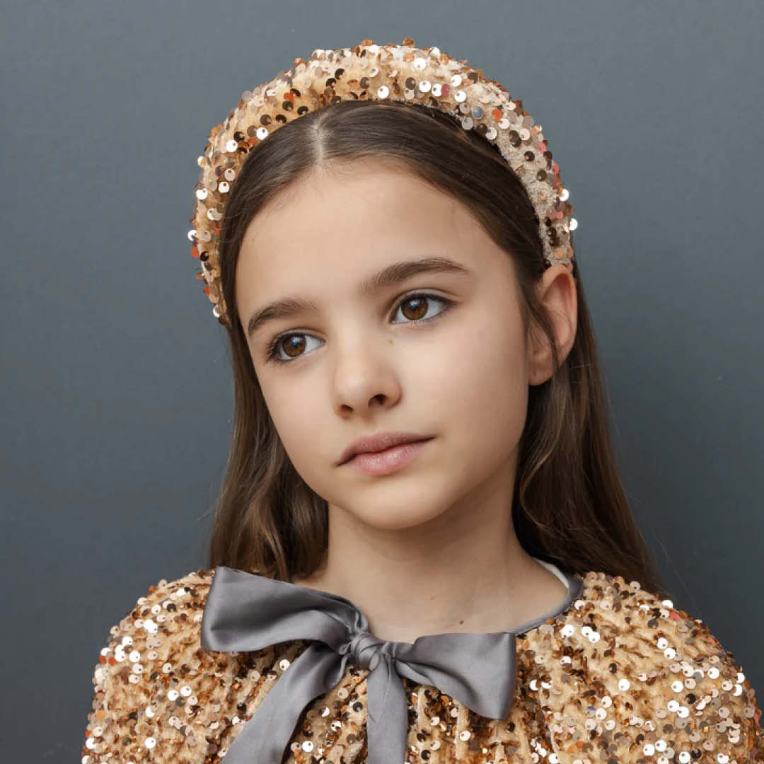 Close-up van een meisje dat de gouden pailletten verkleedcape van Mimi & Lula draagt met een grijze strik en een bijpassende haarband.