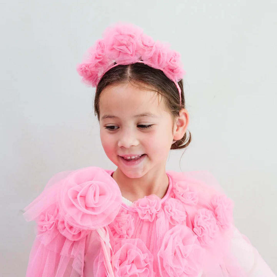 Meisje met de roze verkleedcape en haarband, lachend en een roze bloemenstaf vasthoudend.