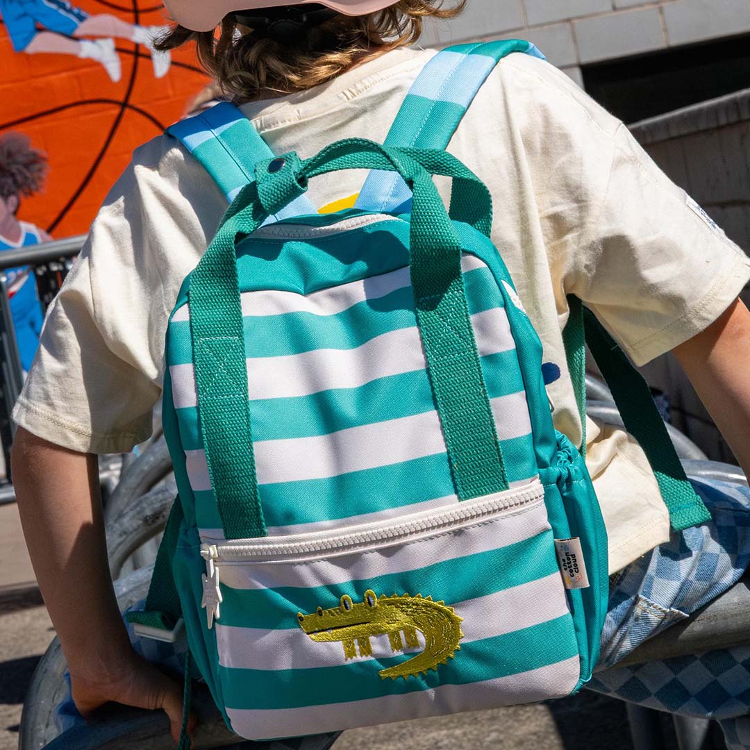 Een kind draagt de the cotton cloud kleuterrugzak met turquoise strepen en krokodil op de rug.