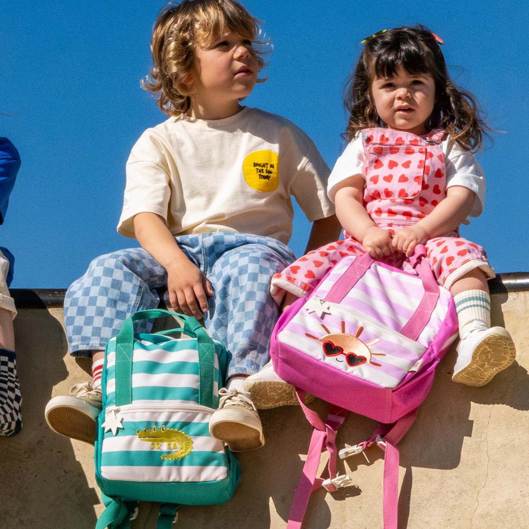 Twee kinderen zitten buiten, één met de the cotton cloud kleuterrugzak met turquoise strepen en krokodil.