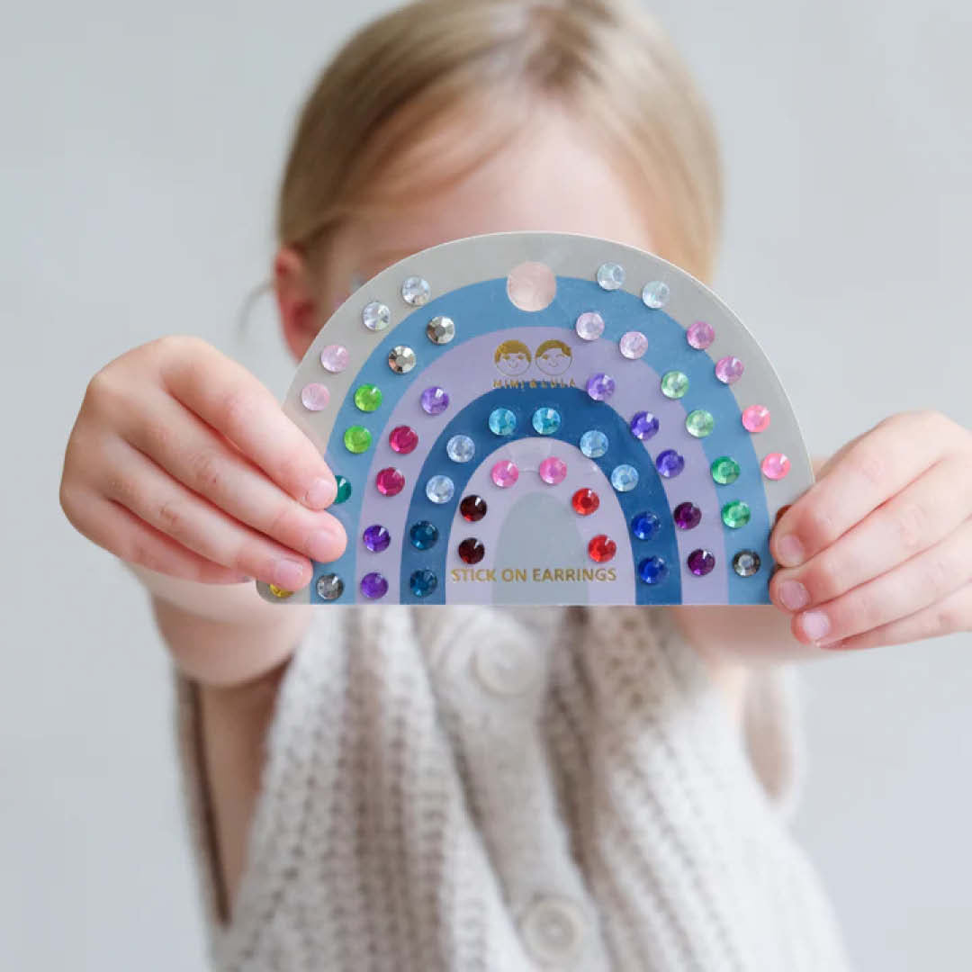Een kind houdt de Mimi & Lula regenboogkaart met veelkleurige plakoorbellen voor zich.