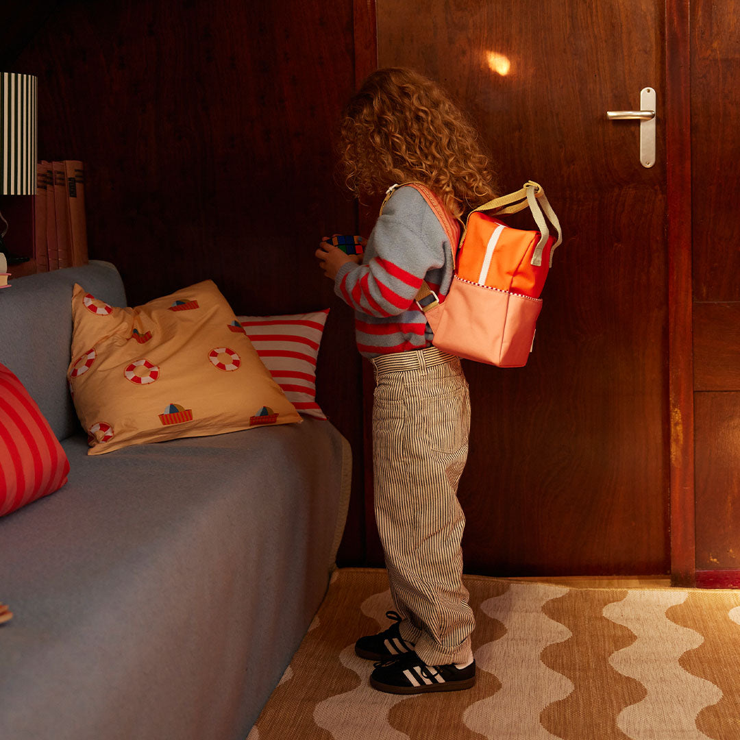 Een kind met krullend haar draagt de Sticky Lemon peuterrugzak in oranje en koraalroze op de rug.