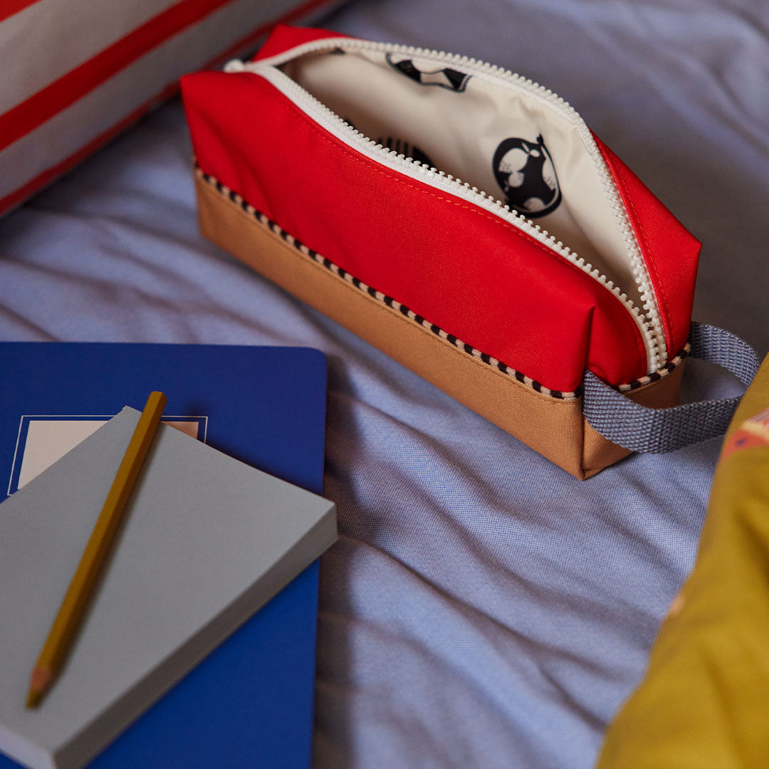 Open colourblocking pennenetui met dierenthema voering op een blauw bed naast een notitieboekje.