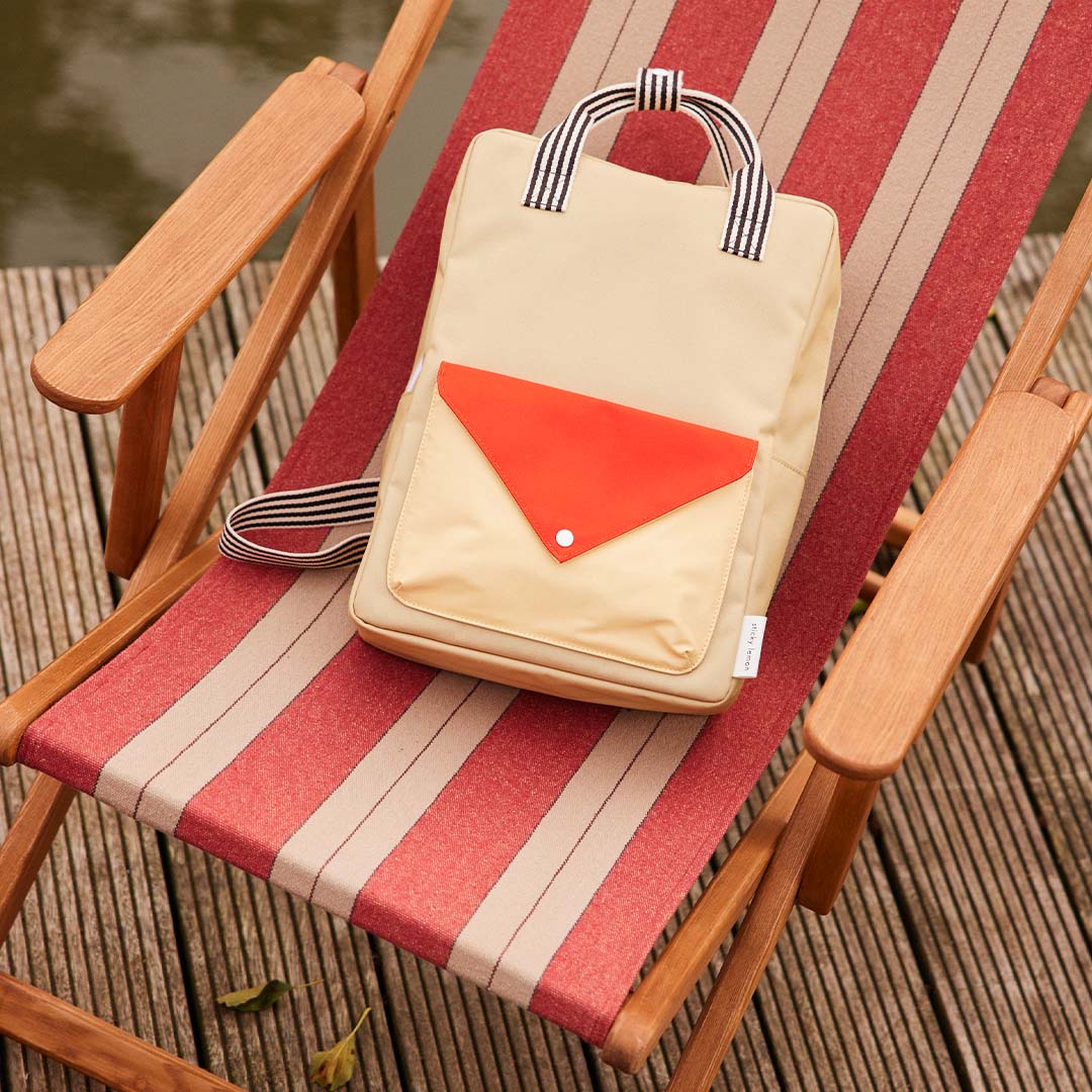Een Sticky Lemon schoolrugzak in beige met rood envelopvak ligt op een gestreepte strandstoel bij het water.