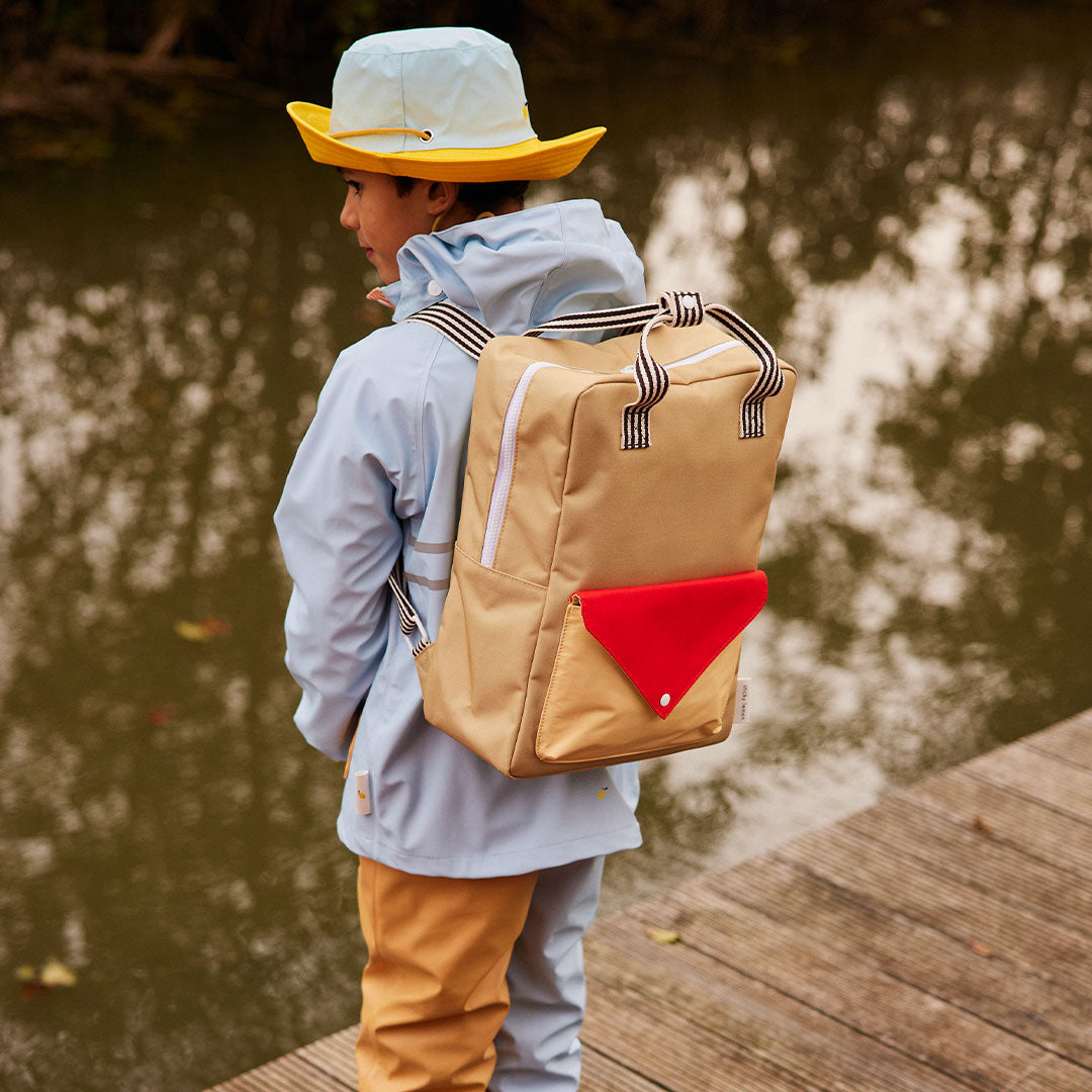 Een kind draagt de Sticky Lemon schoolrugzak in beige met rood envelopvak op een houten vlonder bij het water.