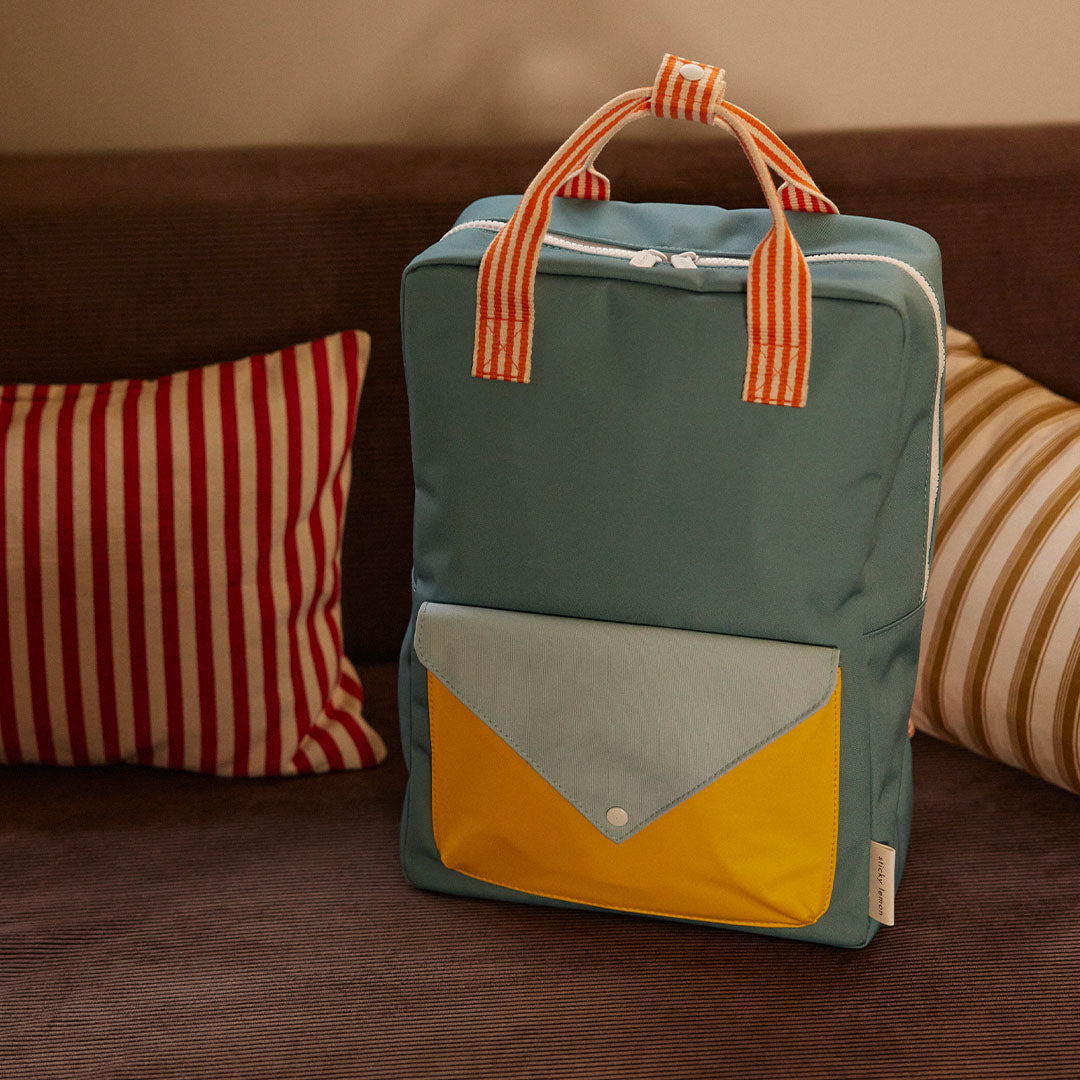 Sticky Lemon schoolrugzak in blauw met envelopzak, staand op een bruine bank met gestreepte kussens.