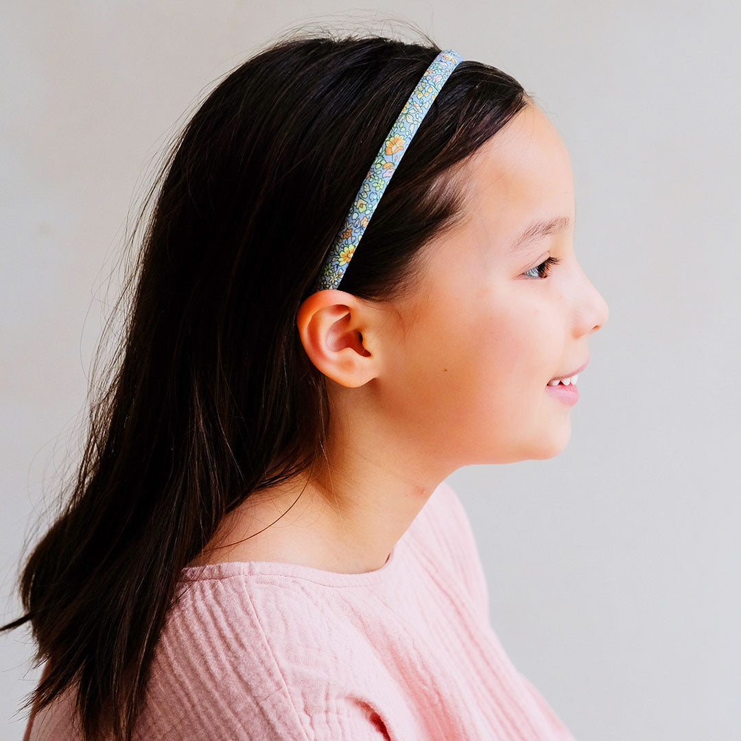 Een kind draagt een mimi & lula gebloemde kinderhoofdband met blauwe achtergrond.