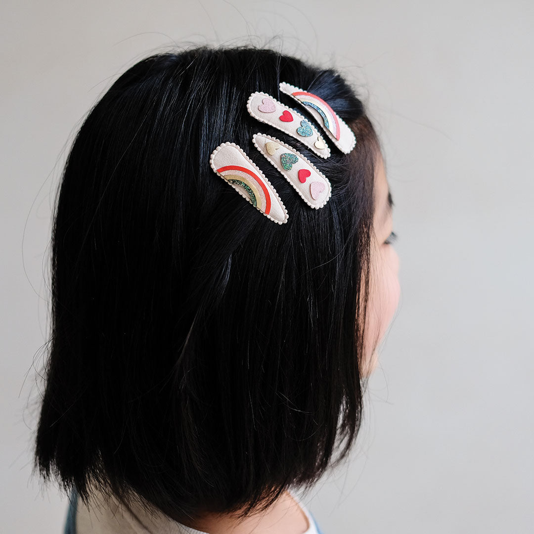 Een meisje met kort zwart haar draagt drie mimi & lula kinder haarclipjes met regenboog- en hartenpatronen.