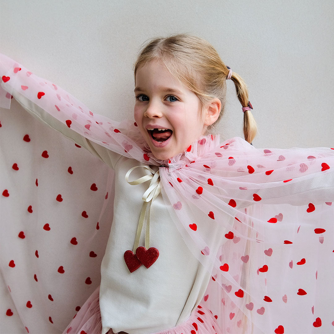 Een lachend meisje draagt de roze kinder verkleedcape met harten en spreidt haar armen.