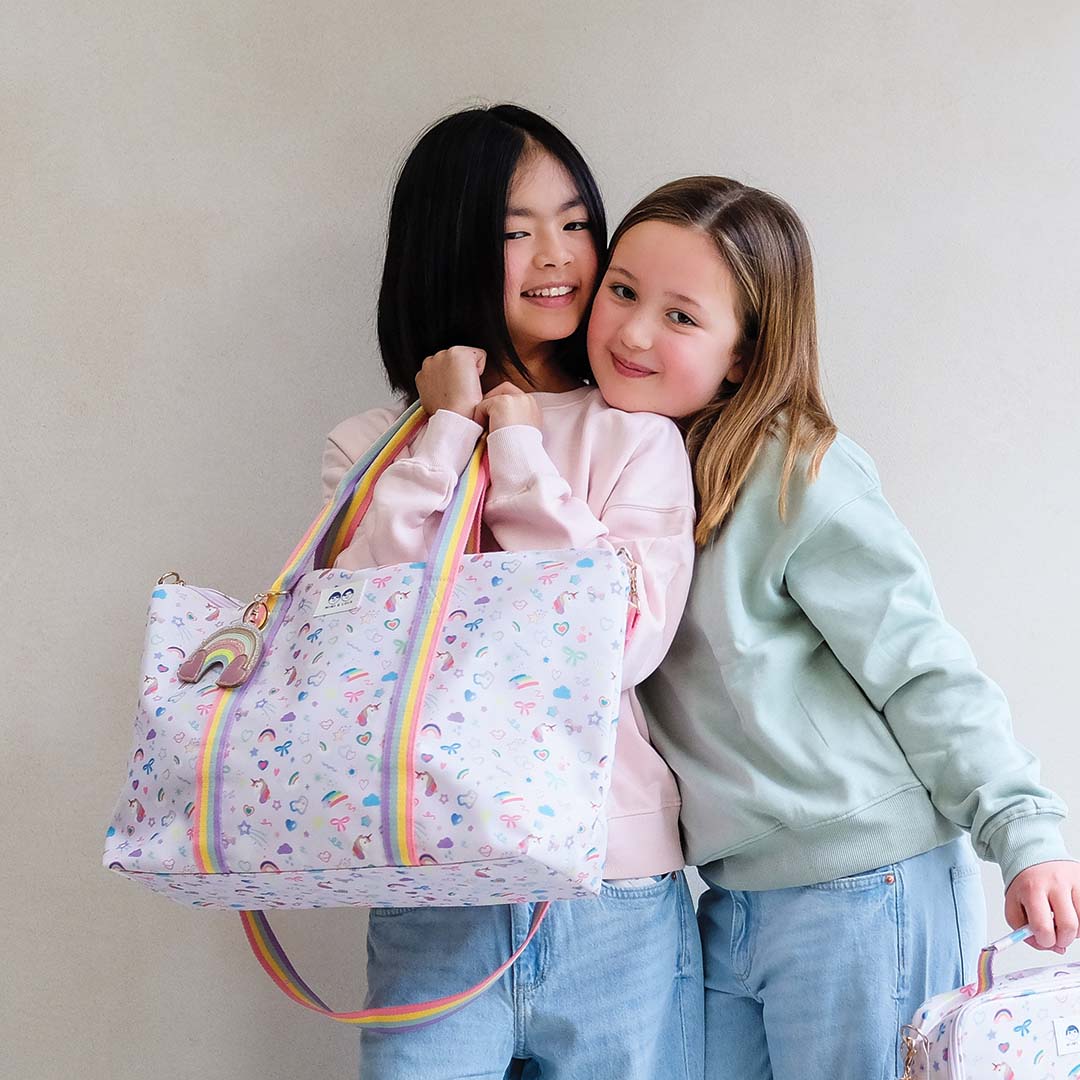Twee lachende meisjes poseren met de Mimi & Lula weekendtas met eenhoorn- en regenboogpatroon.