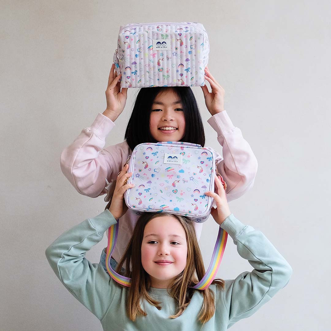 Twee lachende meisjes met een Mimi & Lula lunchtas en een bijpassende tas met eenhoorn- en regenboogpatroon op hun hoofd.
