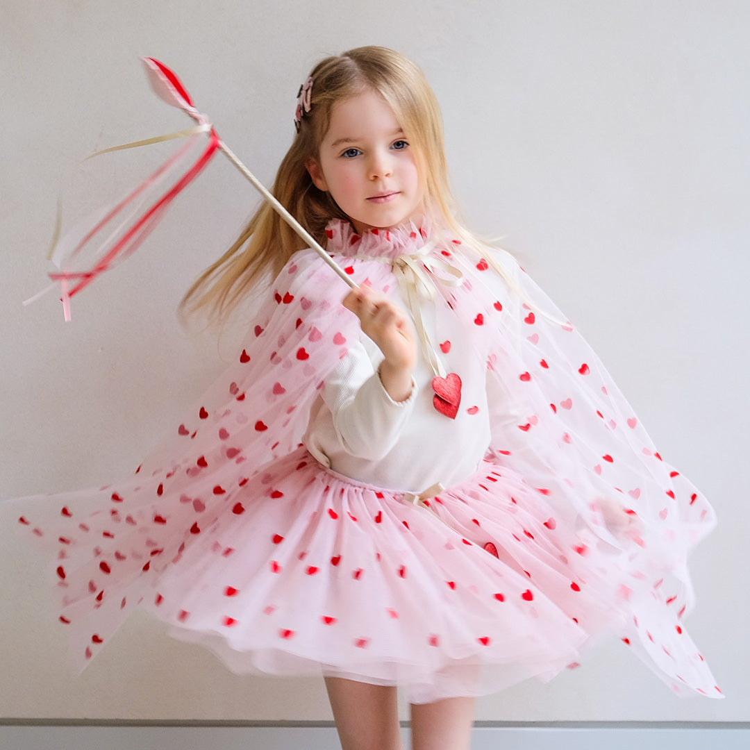 Een jong meisje draagt een mimi & lula roze verkleedrok en cape met rode harten en houdt een hartjesstaf vast.