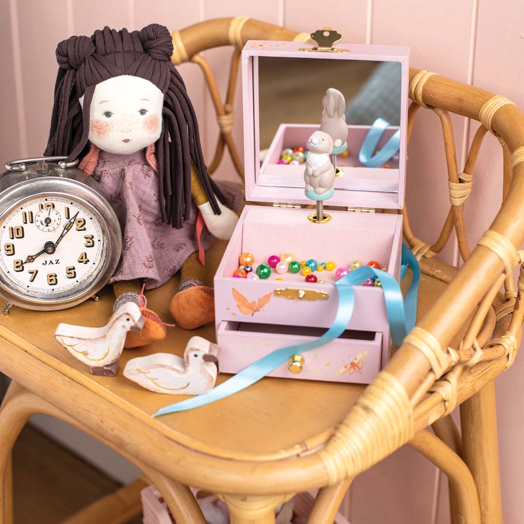Moulin Roty roze muzieksieradendoos open met konijn en kralen, naast een pop en wekker op een rieten stoel.