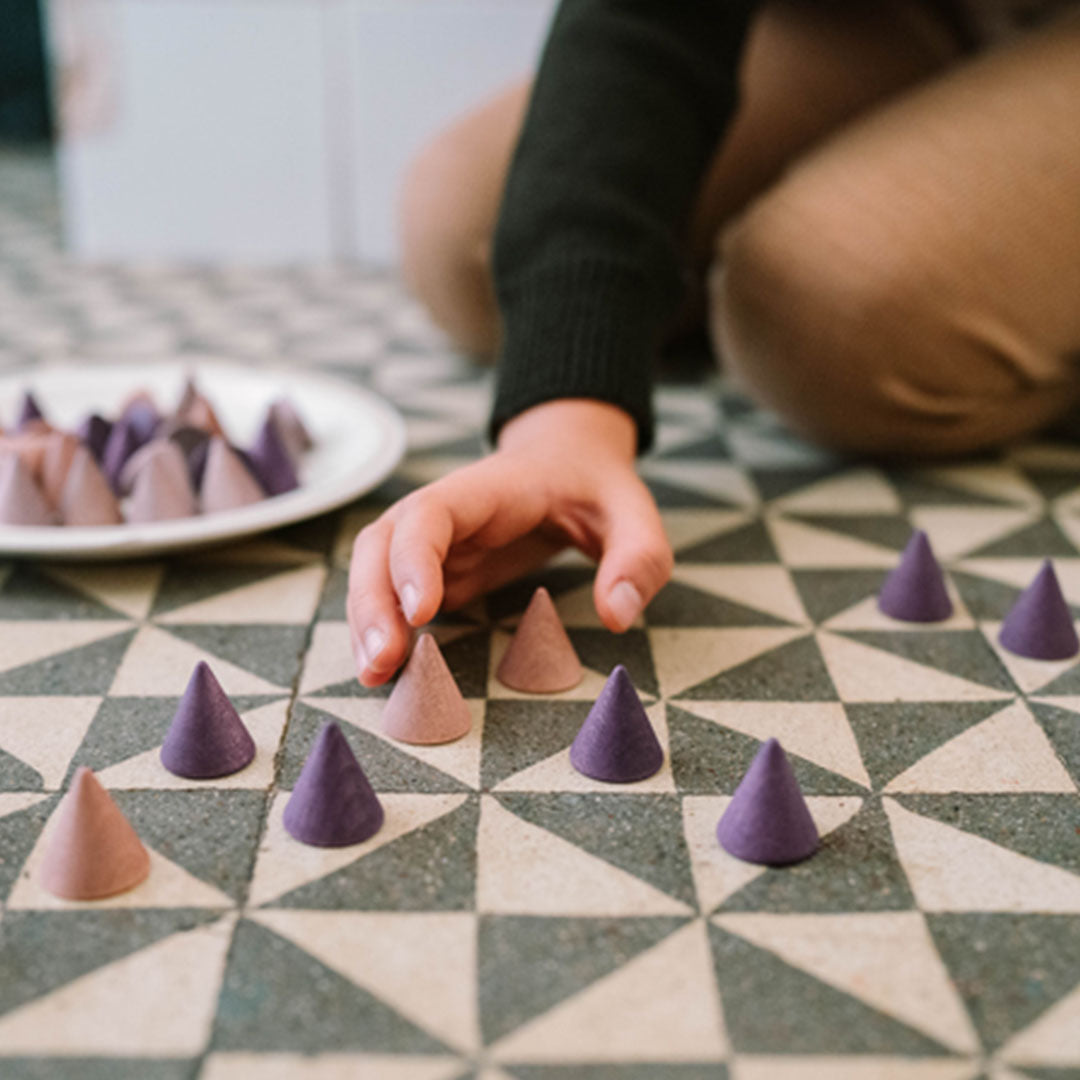 Een kinderhand pakt een grapat houten kegel van een tegelvloer, met meer kegels en een bord op de achtergrond.
