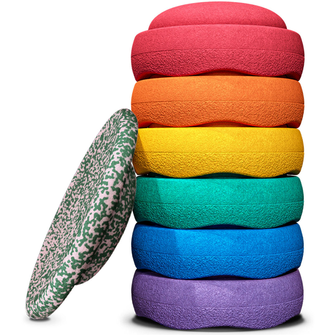 Een stapel van zes ronde stapelstein schuimrubberblokken in regenboogkleuren, met een zevende groen gespikkeld blok ernaast.