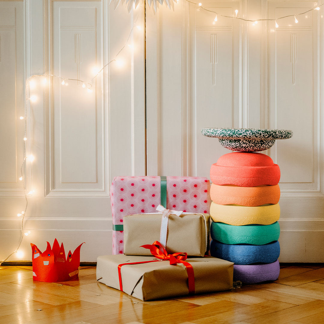 Een stapel van zeven stapelstein schuimrubberblokken in regenboogkleuren staat naast ingepakte cadeaus op een houten vloer.