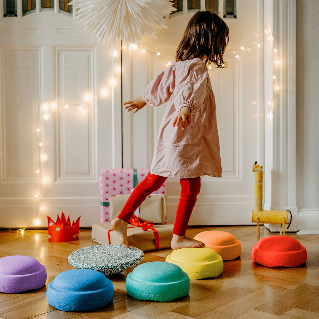 Een meisje balanceert op verspreide stapelstein schuimrubberblokken in regenboogkleuren op een houten vloer.