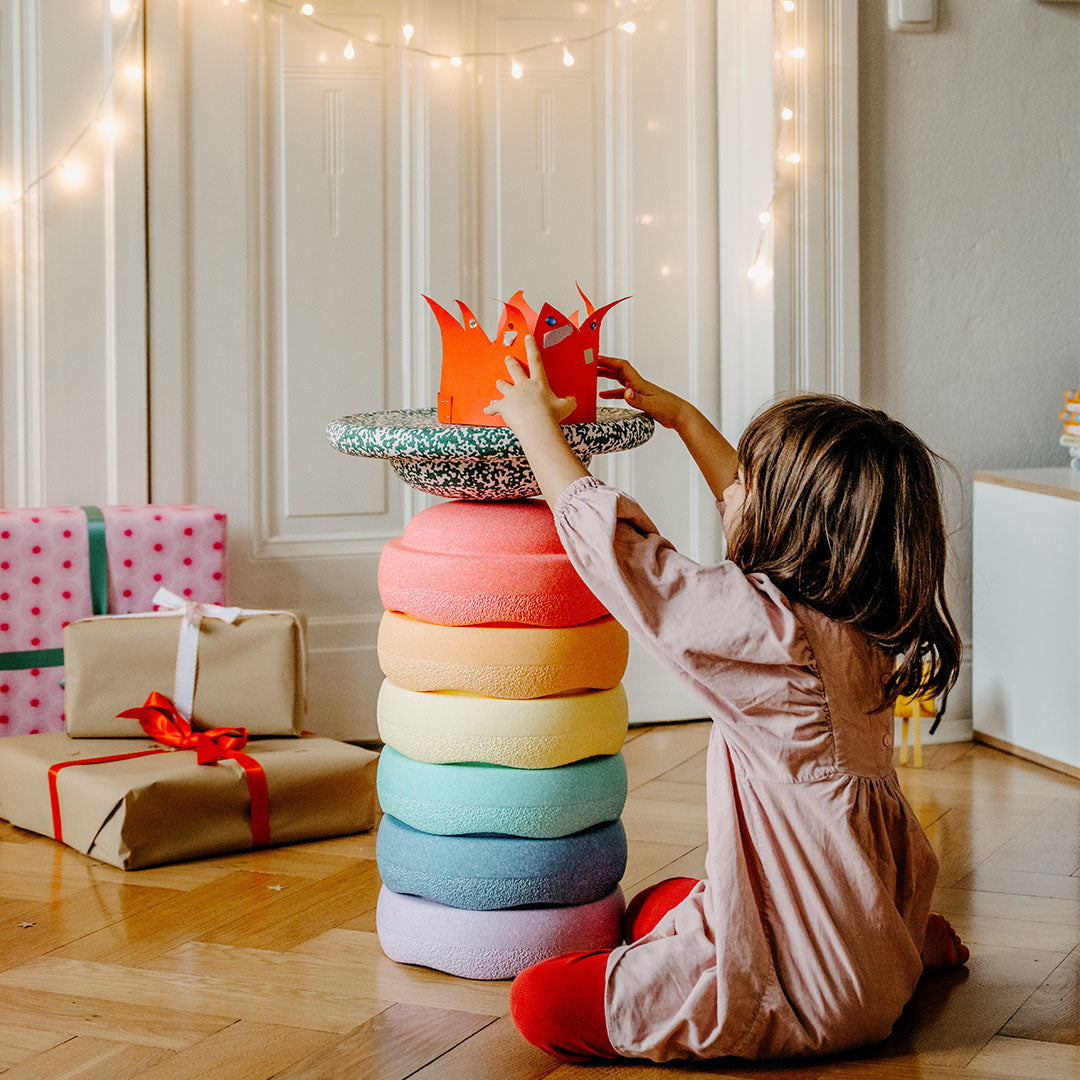 Een kind speelt met stapelstein bouwstenen in pastelkleuren, met cadeaus en kerstverlichting op de achtergrond.