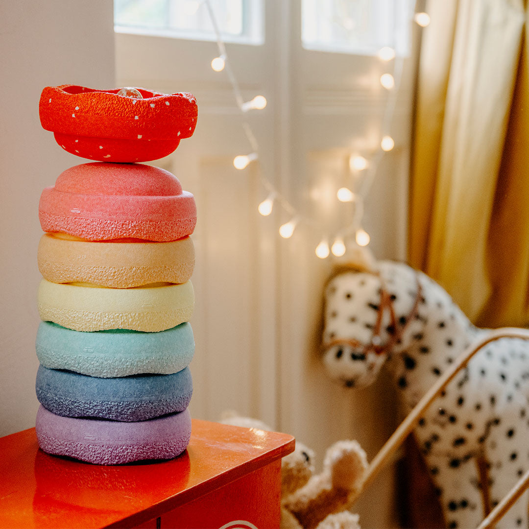 stapelstein stapelspeelgoed van zeven ronde, pastelkleurige stenen met textuur, op een rood kastje, met een hobbelpaard op de achtergrond.