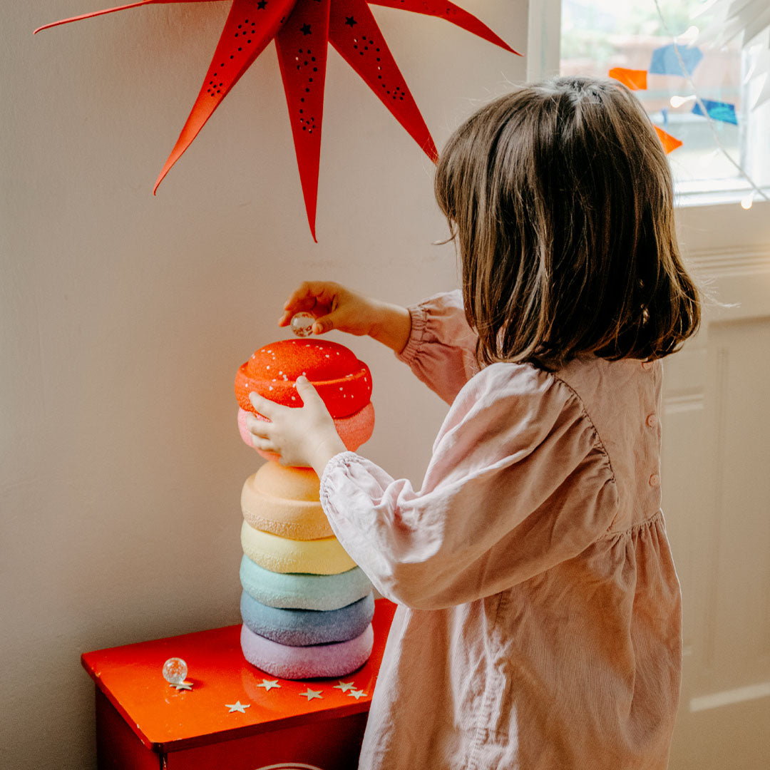 Een kind in een roze jurk stapelt de rode stapelstein steen op een toren van pastelkleurige stenen.