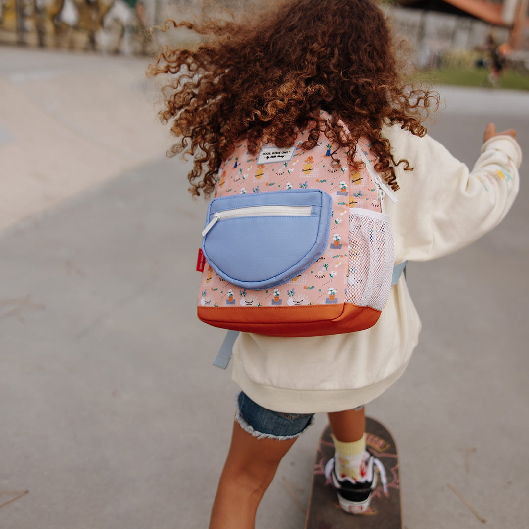 Kind met krullend haar draagt de Hello Hossy schoolrugzak Flore tijdens het skateboarden.