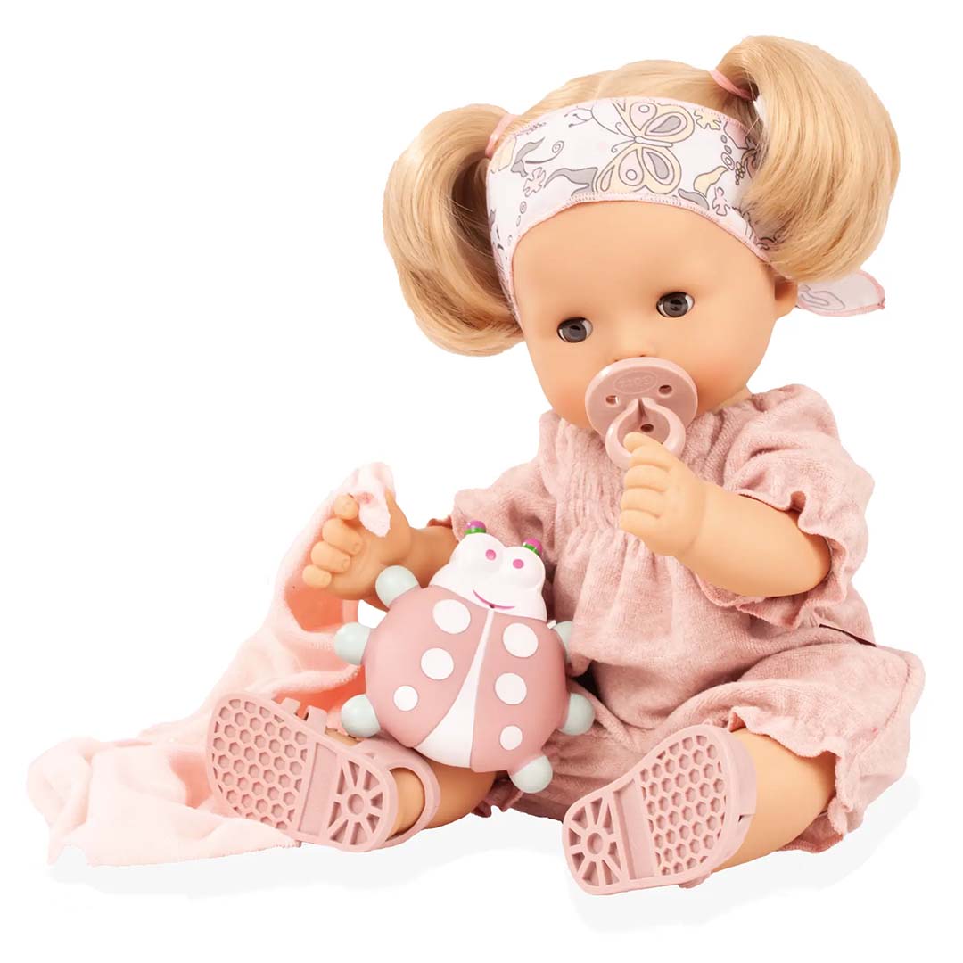 Gotz babypop Maxy Aquini met blonde pigtails, roze kleding en een hoofdband met vlinderpatroon, zittend met een speen, roze deken en lieveheersbeestje.
