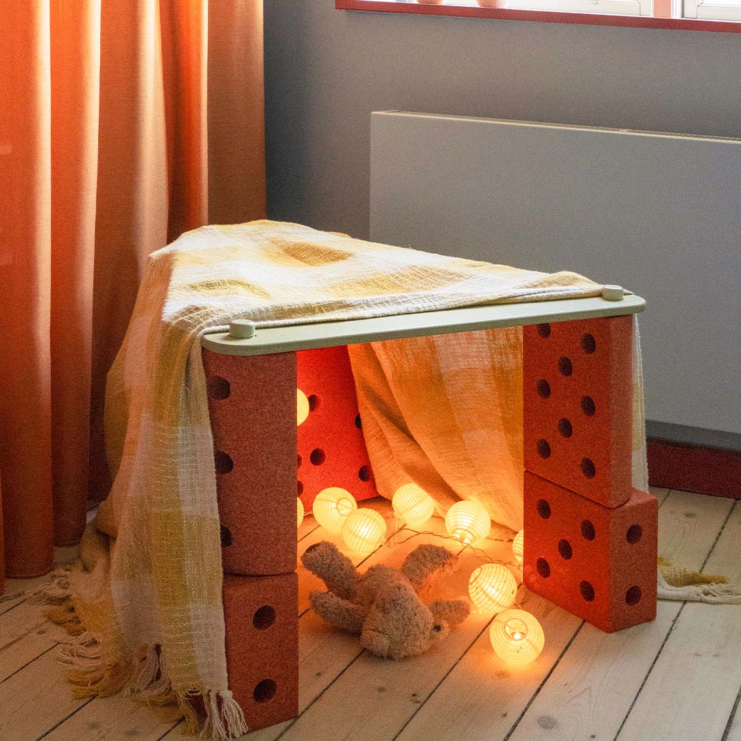 Een speelhuisje gemaakt van modu evenwichtsbalken en oranje bouwblokken, bedekt met een deken en verlicht met lichtslingers.