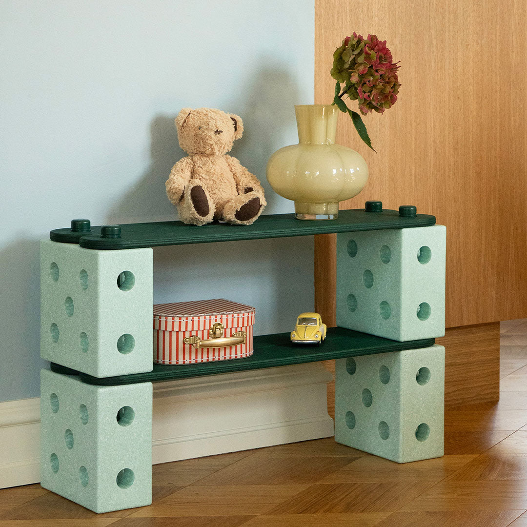Een modu speelgoedmeubel gemaakt van groene houten balken en mintgroene blokken, met een teddybeer en vaas erop.