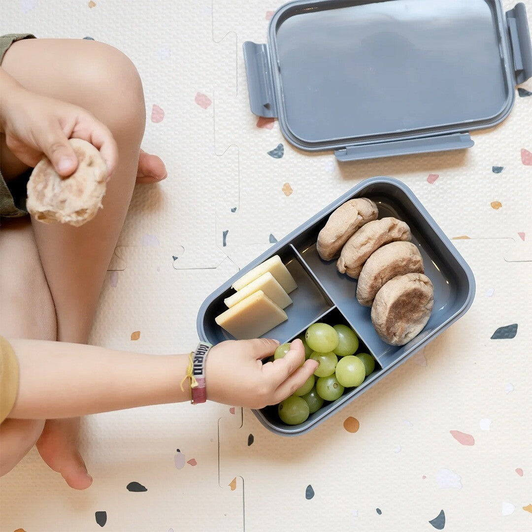 Een kind pakt druiven uit de open 3 sprouts bento kinder lunchbox op een speelmat.