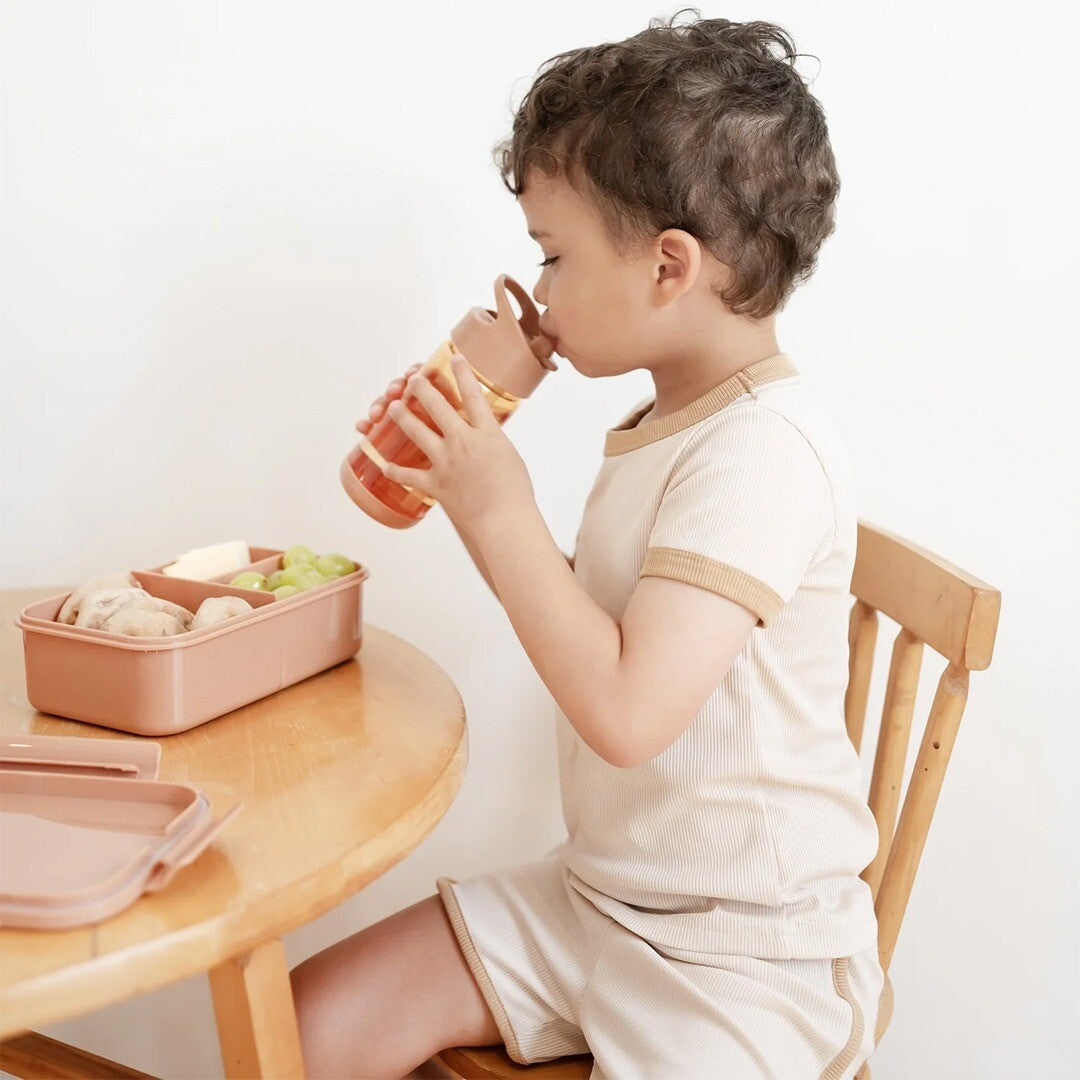 Een kind drinkt uit een flesje naast een open 3 sprouts bento broodtrommel op een houten tafel.