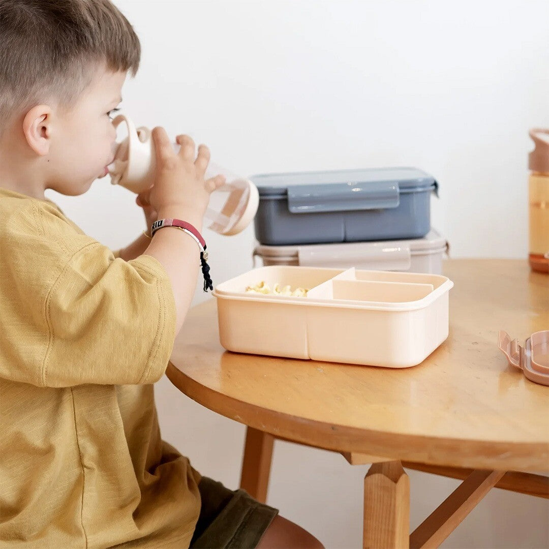 Een kind drinkt uit de 3 sprouts kinder drinkfles aan een houten tafel met lunchboxen.