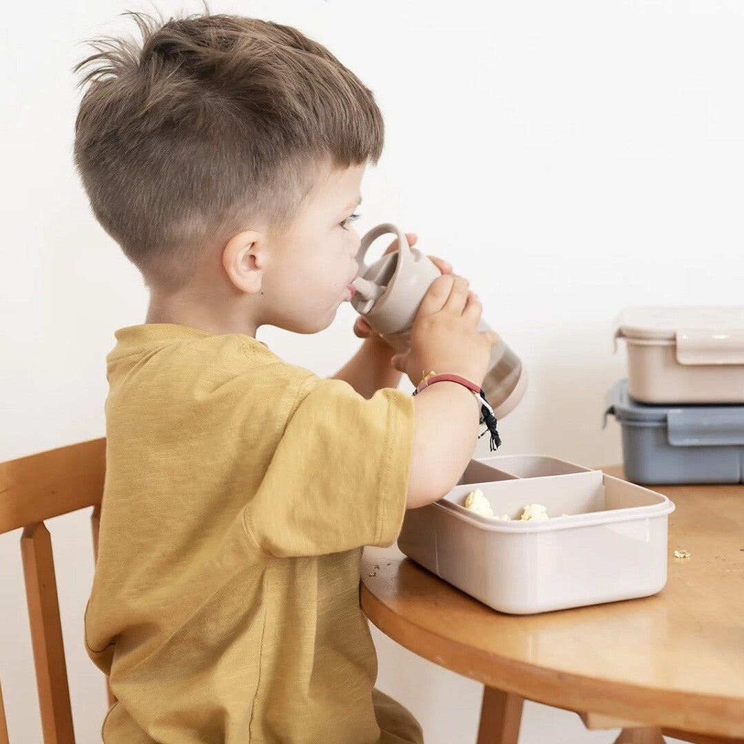 Een kind drinkt uit de 3 sprouts taupe drinkfles aan tafel, met een lunchbox ernaast.