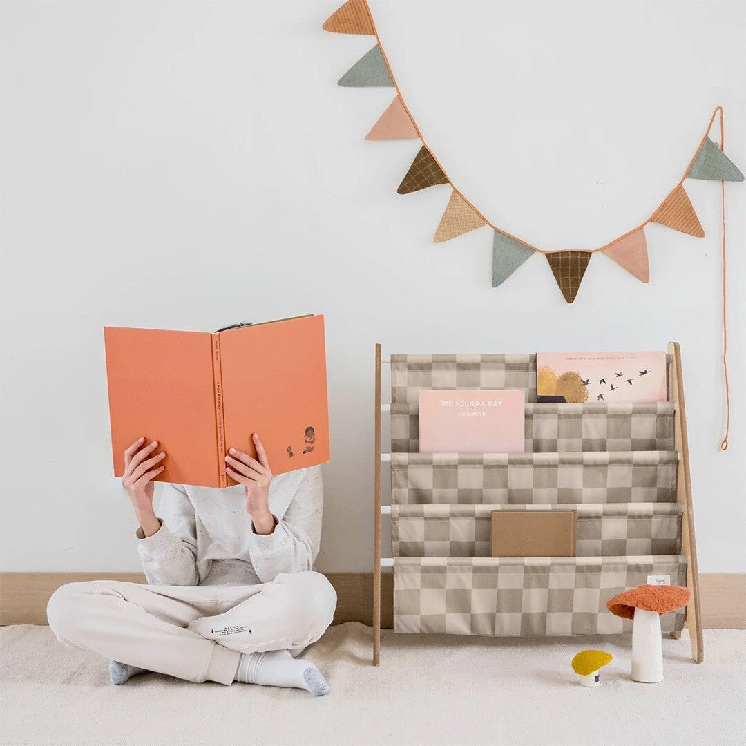 Kind leest een boek naast het 3 sprouts kinderboekenrek met beige geruite stoffen vakken.