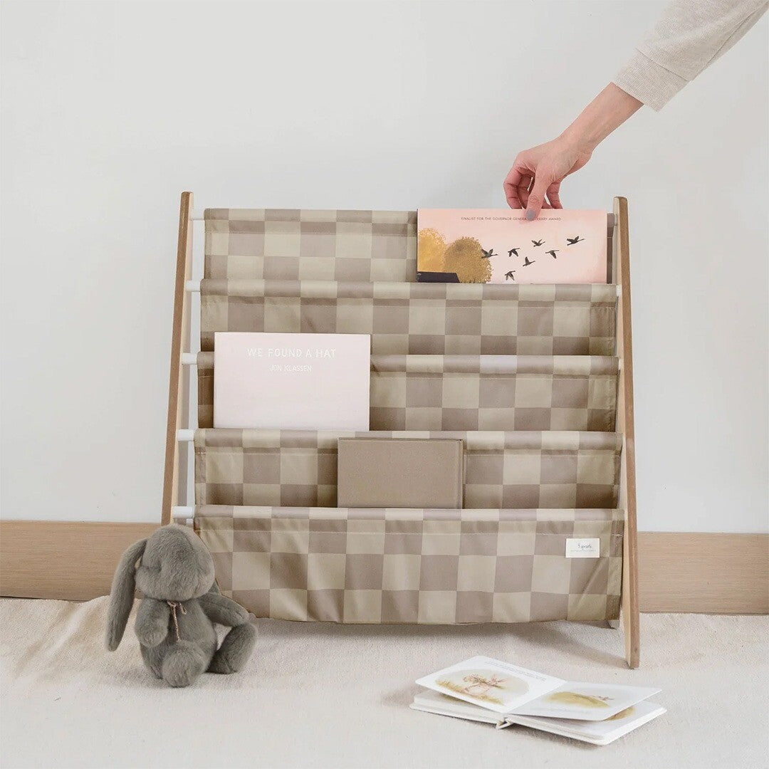 Een hand plaatst een boek in het 3 sprouts kinderboekenrek met beige geruite stoffen vakken.