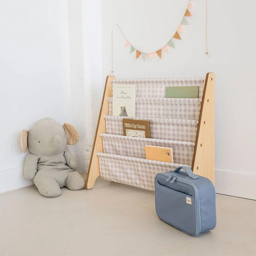 3 sprouts kinderboekenrek met beige gingham patroon, gevuld met boeken, naast een olifantenknuffel en een blauwe tas.