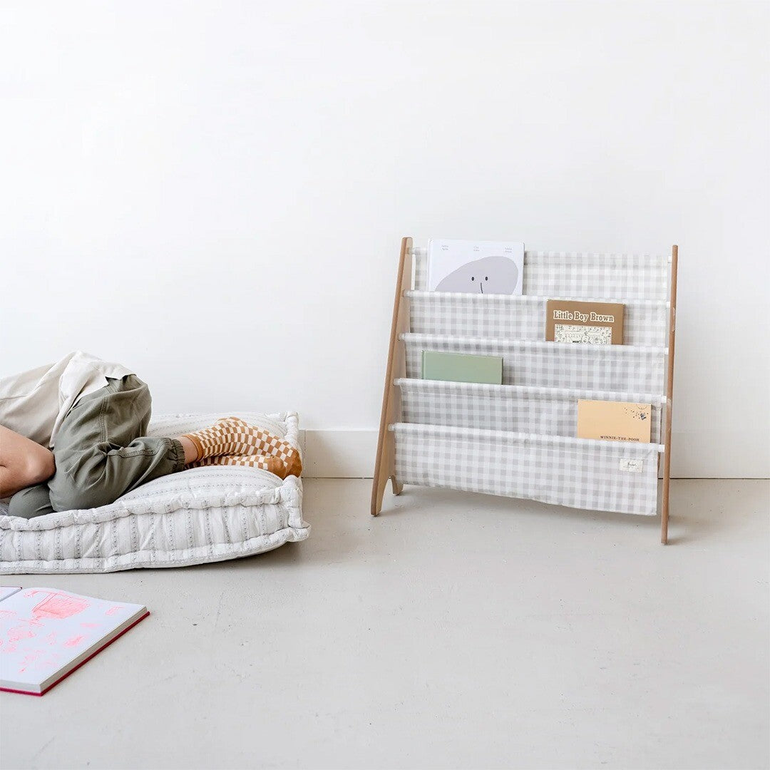 3 sprouts kinderboekenrek met beige gingham patroon, gevuld met boeken, in een kinderkamer met een kind op een kussen.
