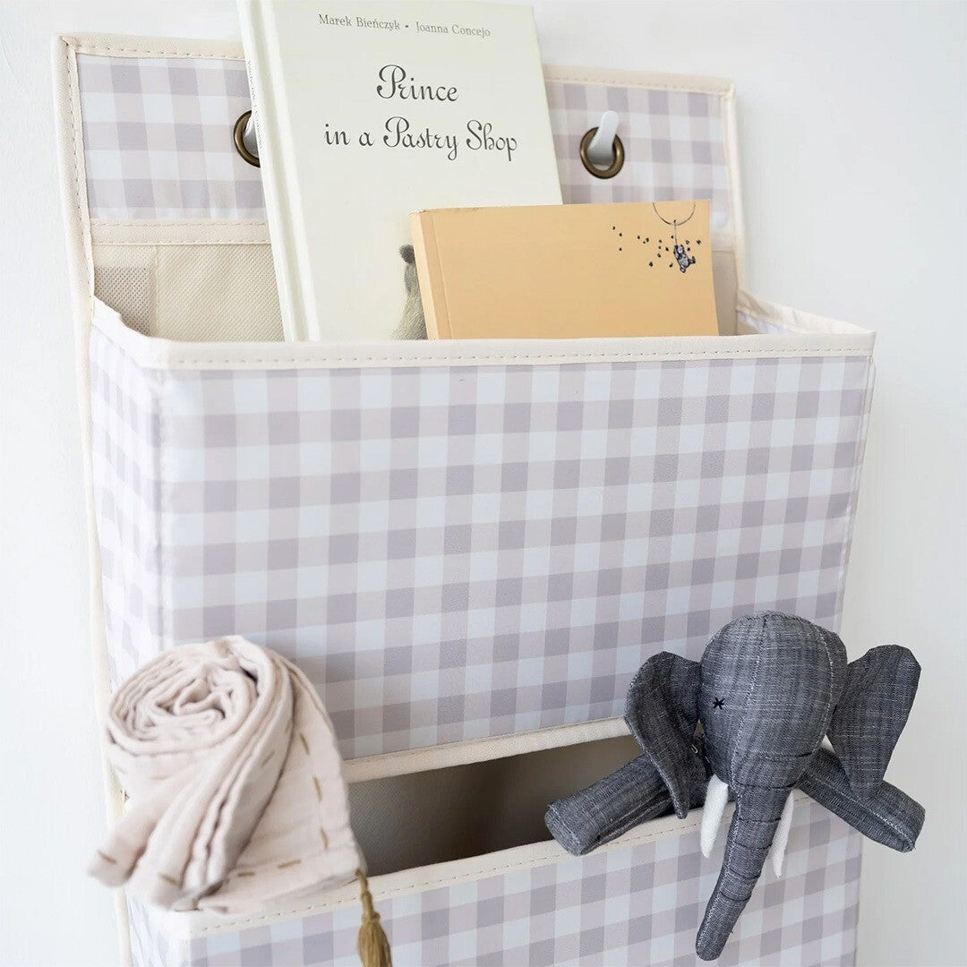 Detail van de beige geruite wandorganiser met een boek, notitieboekje, doek en een grijze olifant knuffel in de vakken.