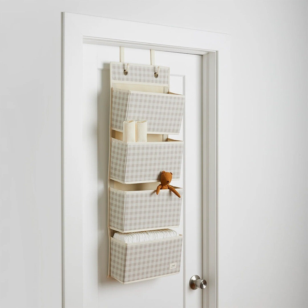 Beige geruite wandorganiser met vier vakken hangend over een witte deur, gevuld met babyspullen en een knuffelbeertje.
