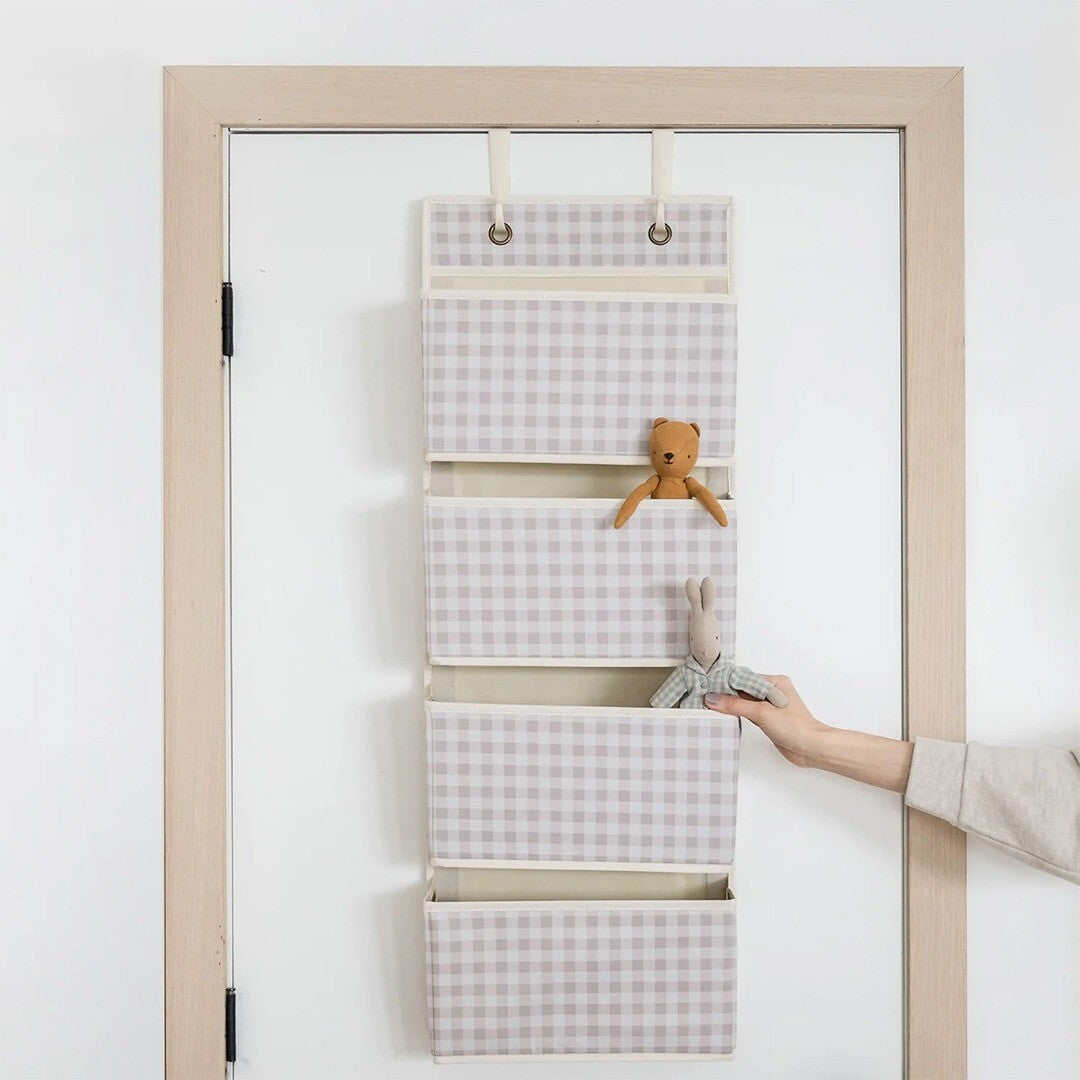 Beige geruite wandorganiser met vijf vakken hangend over een deur, met een hand die een konijn knuffel in een vak plaatst.