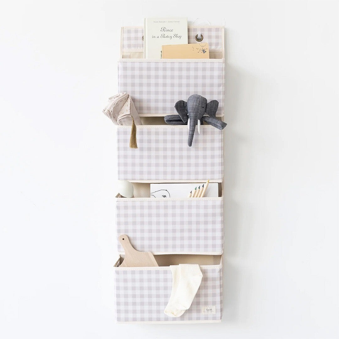 Beige geruite wandorganiser met vijf vakken hangend aan een muur, gevuld met een boek, olifant knuffel, potloden en andere spullen.